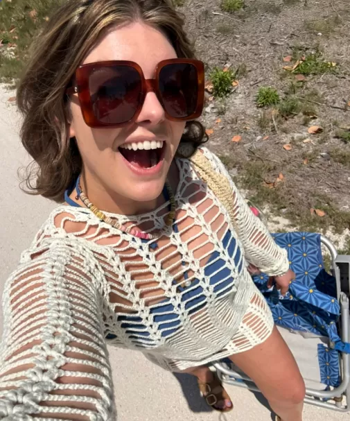Woman in oversized sunglasses and crochet cover-up holding a beach chair.